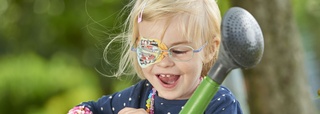 Kopfgrafik lachendes Kind mit Feuerwehr-Augenpflaster und Gießkanne in der Hand